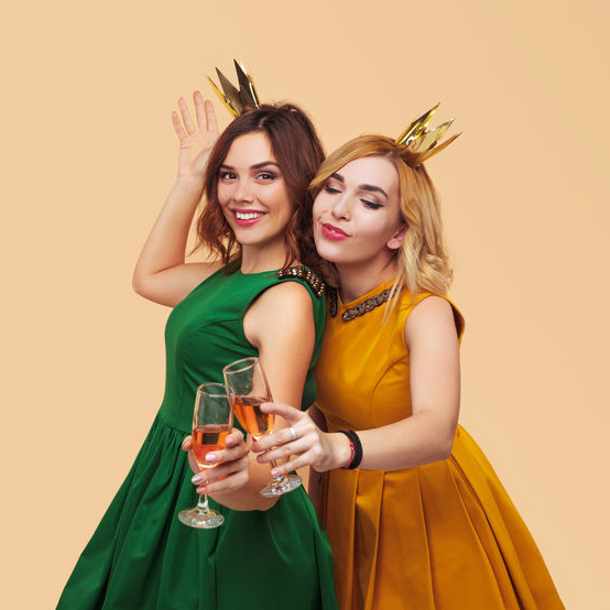 Cheerful content women in stylish colorful dresses and golden crowns celebrating New Year party clinking with champagne glasses on beige background